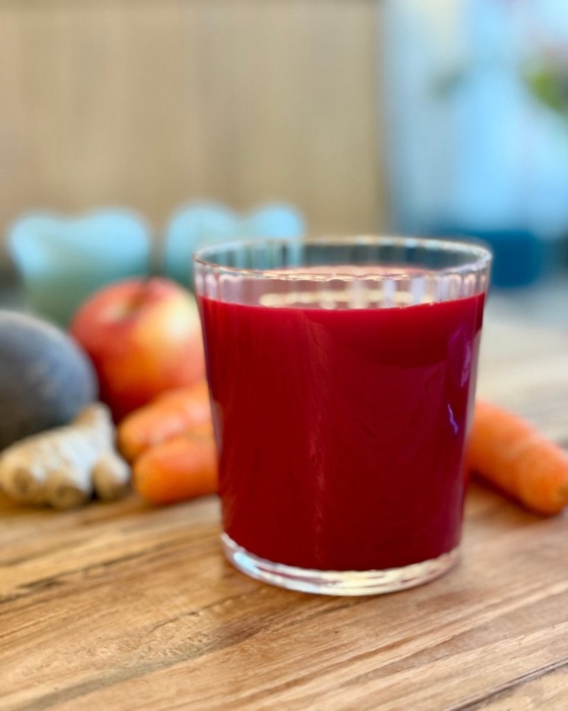 A glass of freshly pressed beet juice, deep red, with carrots in the background. Beet juice widens blood vessels and improves how well oxygen reaches the skin — the driver behind the warm, lit-from-within quality of the raw food glow.