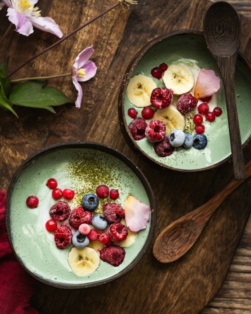 Two green smoothie bowls loaded with fresh raspberries, blueberries, banana, and red currants — the kind of antioxidant-rich raw food combination that defends collagen and builds the raw food glow from within.  