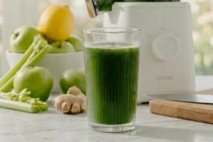 A wide panoramic view of a white Nama cold press juicer and a glass of vibrant green juice on a marble counter, surrounded by an abundant harvest of green apples, celery, ginger, and lemons.