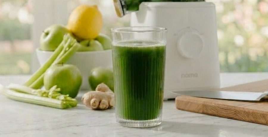 A wide panoramic view of a white Nama cold press juicer and a glass of vibrant green juice on a marble counter, surrounded by an abundant harvest of green apples, celery, ginger, and lemons.
