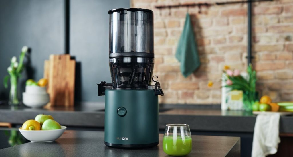 Hurom H320N cold press juicer on a dark kitchen counter with a glass of fresh green juice and green apples.