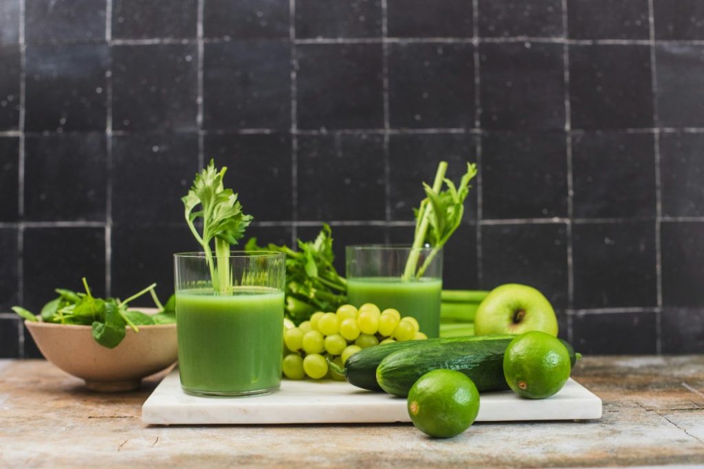 Celery juice on a kitchen countertop surrounded by ingredients such as spinach, celery, apples, cucumber, limes and grapes.