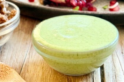 A small glass bowl of creamy, bright green cilantro lime dressing made with hemp seeds, sitting on a wooden table next to a bowl of crumbled walnut meat.