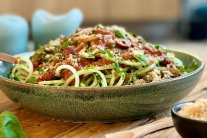 A green ceramic bowl of raw pasta puttanesca — cucumber noodles topped with rich tomato-olive sauce, fresh basil, parsley and macadamia parmesan — on a light wood surface