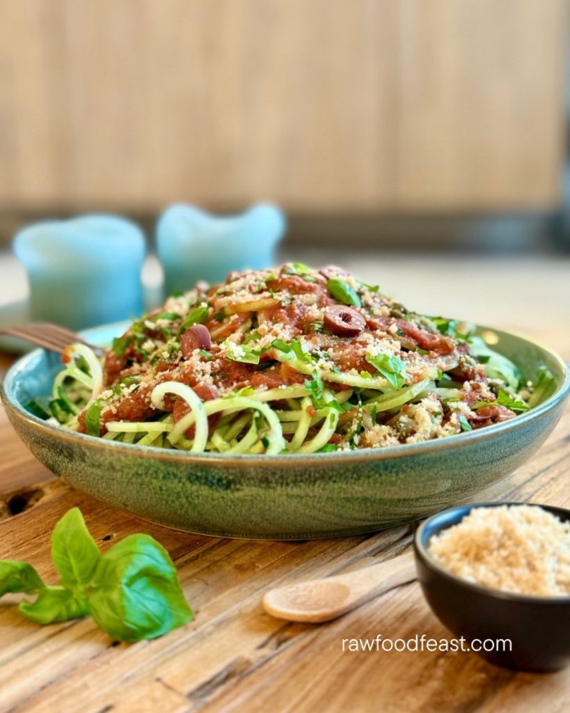 Raw pasta puttanesca in a green ceramic bowl — cucumber noodles with tomato-olive-caper sauce, fresh basil, parsley, and macadamia parmesan on a light wood surface