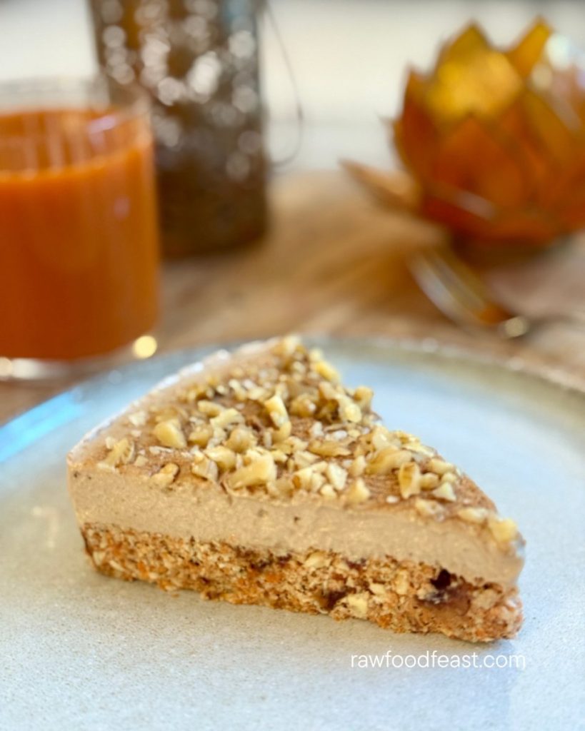 A slice of raw carrot pulp cake with coconut caramel cashew frosting and chopped walnuts on a ceramic plate, served alongside a glass of fresh carrot juice.