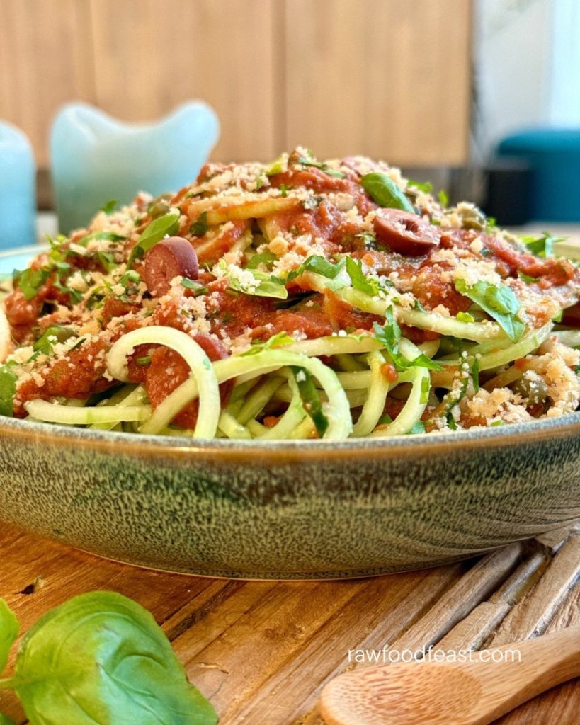 Close-up of raw pasta puttanesca — cucumber noodles loaded with tomato sauce, kalamata olives, capers, fresh basil, parsley, and macadamia parmesan