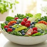 A vibrant, sun-drenched flat lay of a tall cold-pressed green juice next to a massive bowl of raw leafy greens and whole berries on a white marble table with a lush garden background.