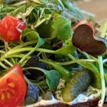 Almond carrot pulp flatbread topped with fresh sprouts, avocado, cherry tomatoes, cornichons, and herbs on a wooden cutting board
