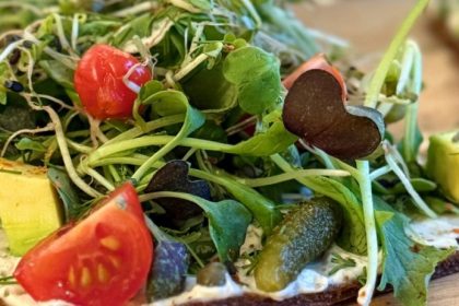 Almond carrot pulp flatbread topped with fresh sprouts, avocado, cherry tomatoes, cornichons, and herbs on a wooden cutting board