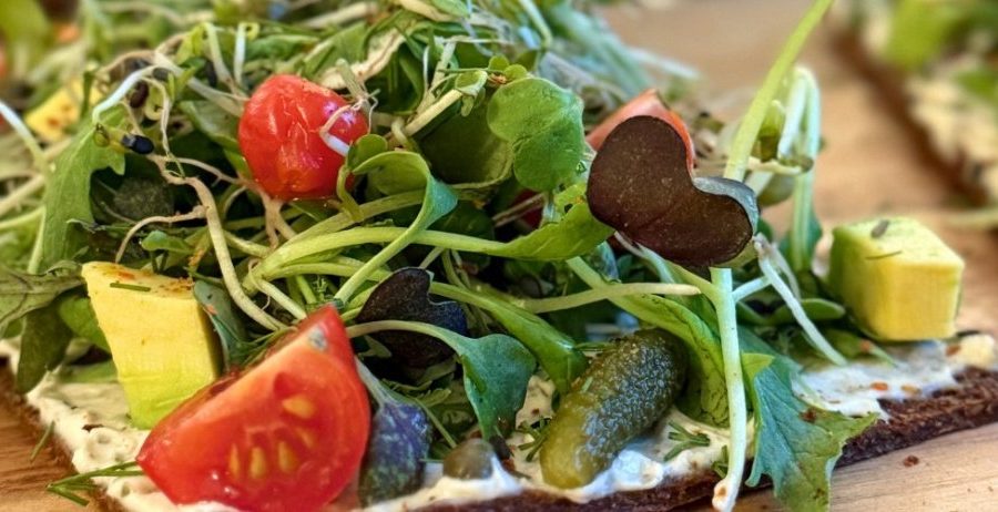 Almond carrot pulp flatbread topped with fresh sprouts, avocado, cherry tomatoes, cornichons, and herbs on a wooden cutting board