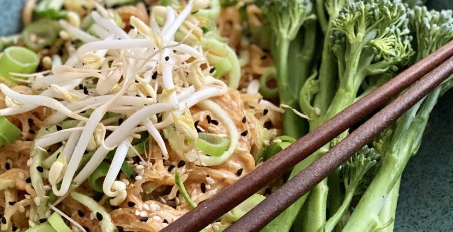 Raw Dan Dan noodles in a teal bowl with kelp noodles, bean sprouts, and fresh broccolini on a light wood surface