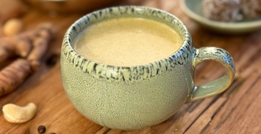 Golden milk recipe in a ceramic mug with fresh turmeric root and cashews on a wooden surface