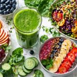 Juicing for weight loss — fresh green juice with colorful fruit bowls, a raw salad with sprouts and dressing, mango, berries, ginger, and leafy greens on a clean white surface