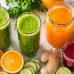 Batch juicing — fresh green juice, orange juice, and beet juice with celery, oranges, apples, cucumber, ginger, and beets on a clean white surface