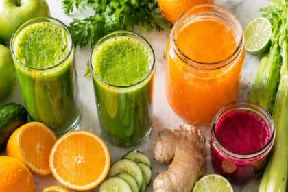 Batch juicing — fresh green juice, orange juice, and beet juice with celery, oranges, apples, cucumber, ginger, and beets on a clean white surface
