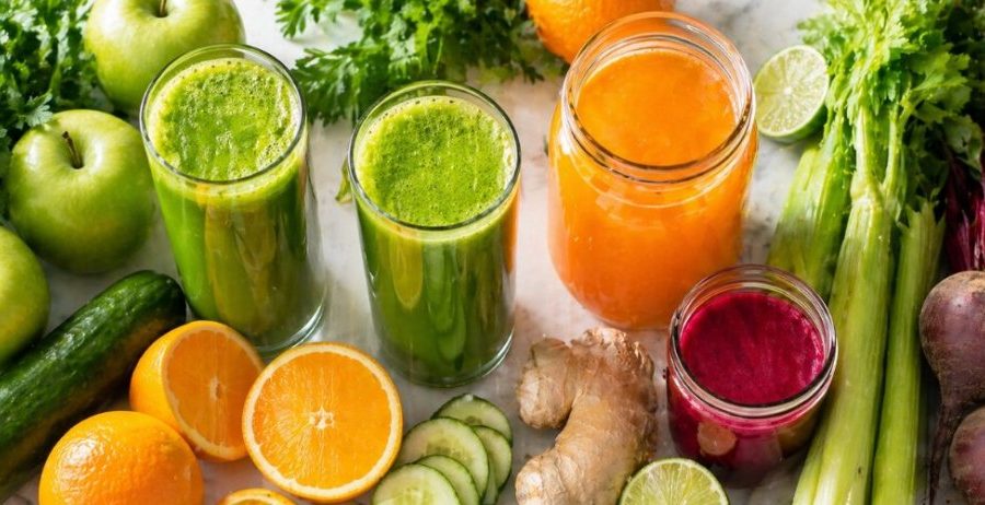 Batch juicing — fresh green juice, orange juice, and beet juice with celery, oranges, apples, cucumber, ginger, and beets on a clean white surface