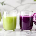 Whole green cabbage and halved red cabbage with two glasses of cabbage juice on white marble — cabbage juice for gut health