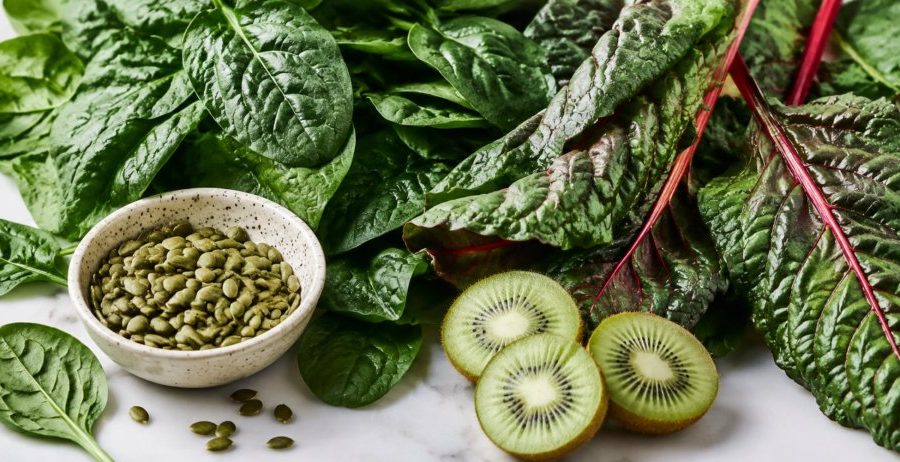 Spinach, swiss chard, pumpkin seeds, and fresh kiwi on white marble — foods for gut health and sleep.