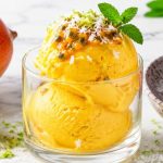 A glass of bright golden mango lime sorbet topped with passion fruit, coconut shred, lime zest, and mint on white marble.