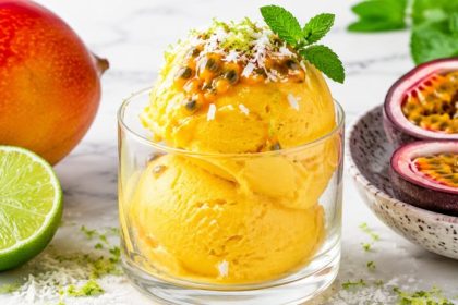 A glass of bright golden mango lime sorbet topped with passion fruit, coconut shred, lime zest, and mint on white marble.
