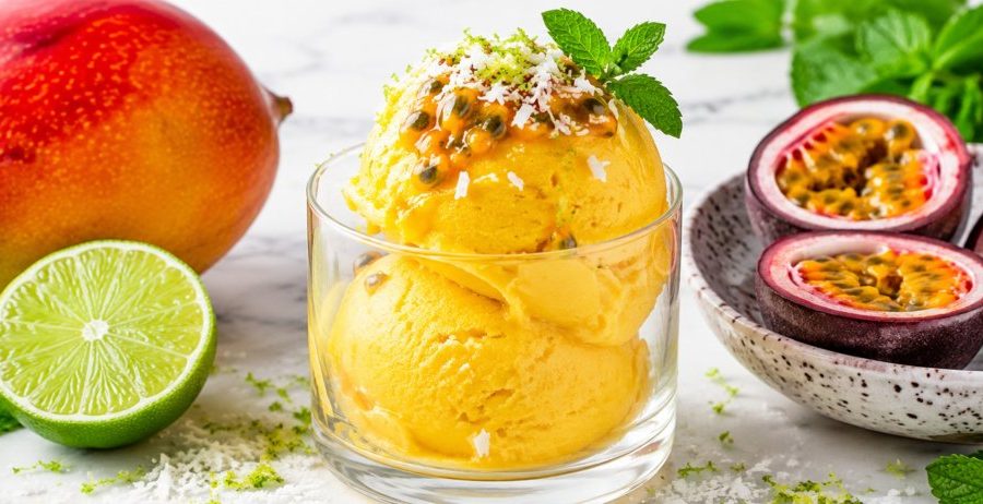 A glass of bright golden mango lime sorbet topped with passion fruit, coconut shred, lime zest, and mint on white marble.