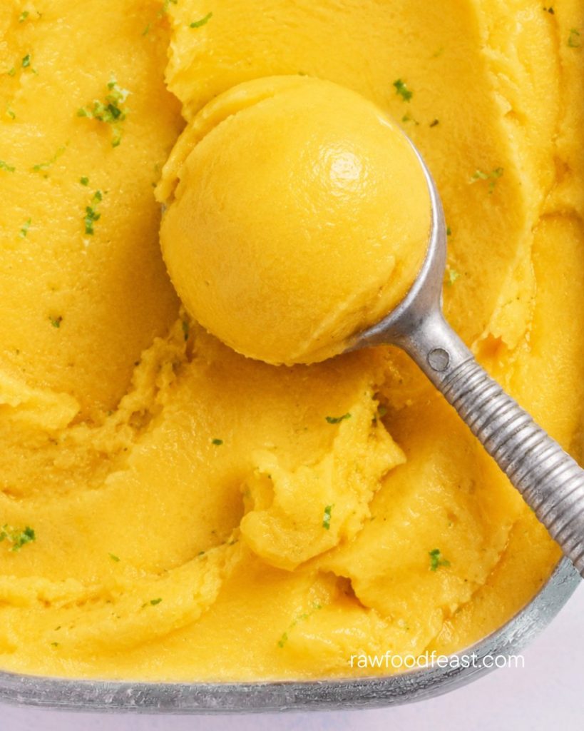 Overhead view of a glass dish of golden mango lime sorbet with an ice cream scoop lifting one perfect round scoop.
