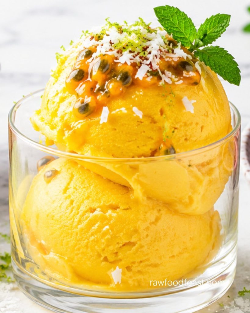Close-up of two scoops of golden mango lime sorbet in a glass topped with passion fruit, coconut shred,
lime zest, and mint.