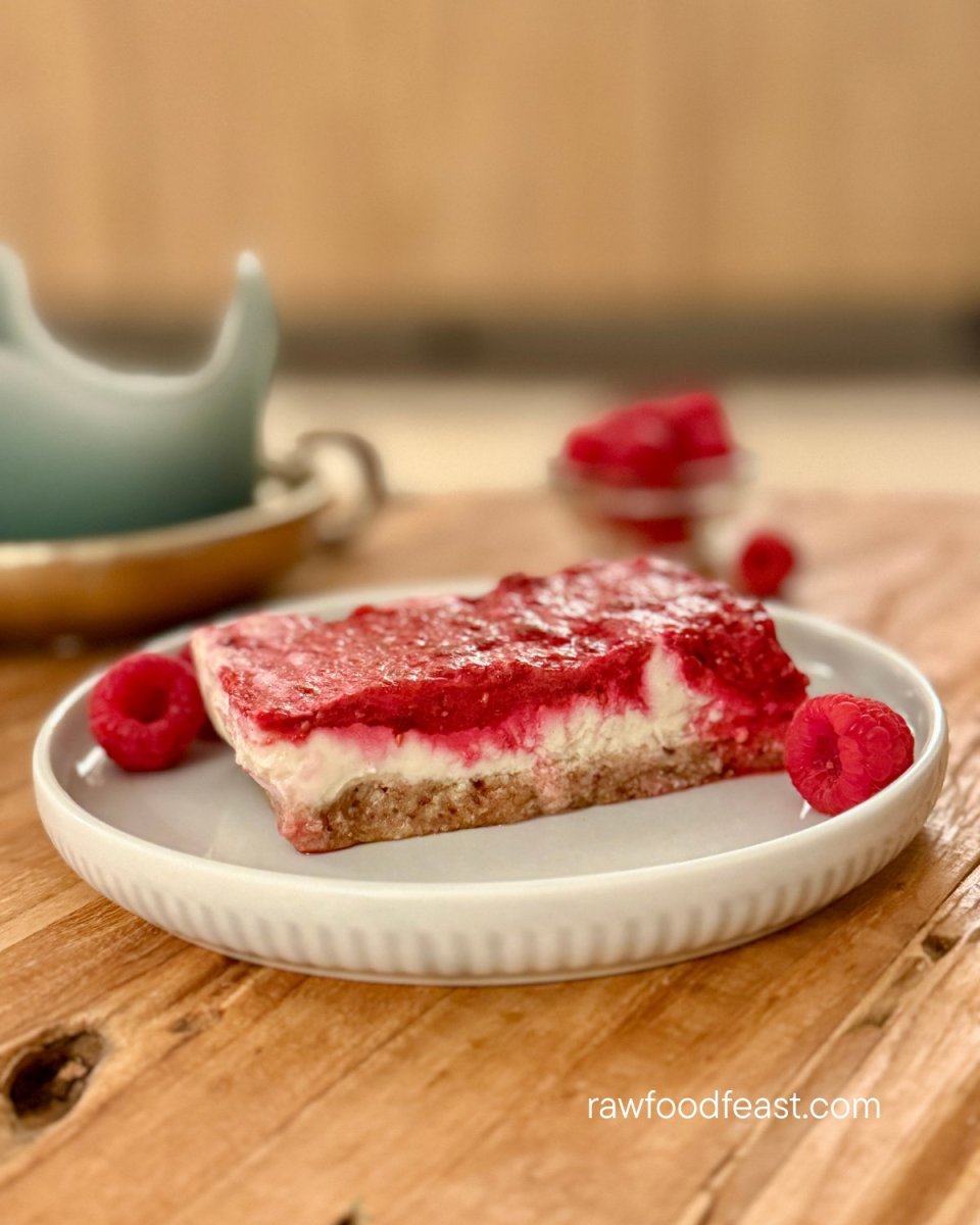 A slice of no bake lemon raspberry bars showing three layers — date crust, creamy lemon filling, and raspberry — with fresh raspberries on a wooden surface