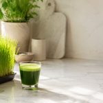 A tray of fresh homegrown wheatgrass beside a shot of bright green wheatgrass juice — the daily wheatgrass juice benefits in one frame.