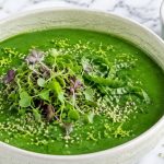 A cream crackle bowl of green gazpacho topped with microgreens, basil, and lime zest on marble — green glow gazpacho.