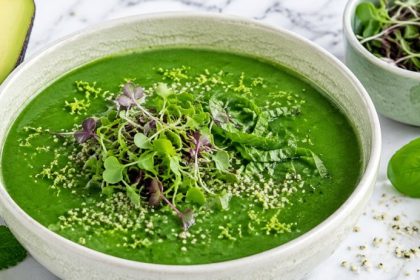 A cream crackle bowl of green gazpacho topped with microgreens, basil, and lime zest on marble — green glow gazpacho.
